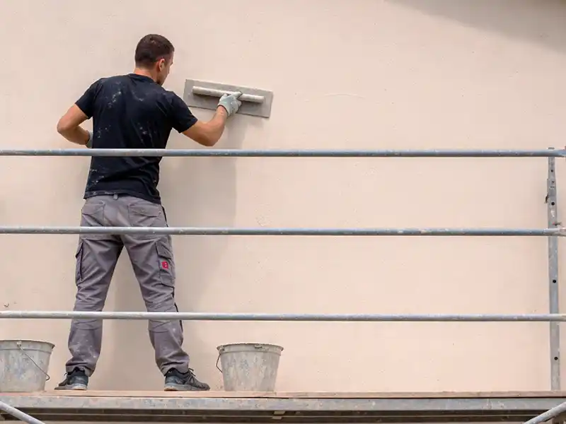 Application enduit de facade pres de Montpellier - Couverture 34 Un homme debout sur un échafaudage effectue l'application d'un enduit de façade, lissant le mur à l'aide d'un outil de plâtrage. Deux seaux métalliques reposent près de ses pieds sur la plate-forme.