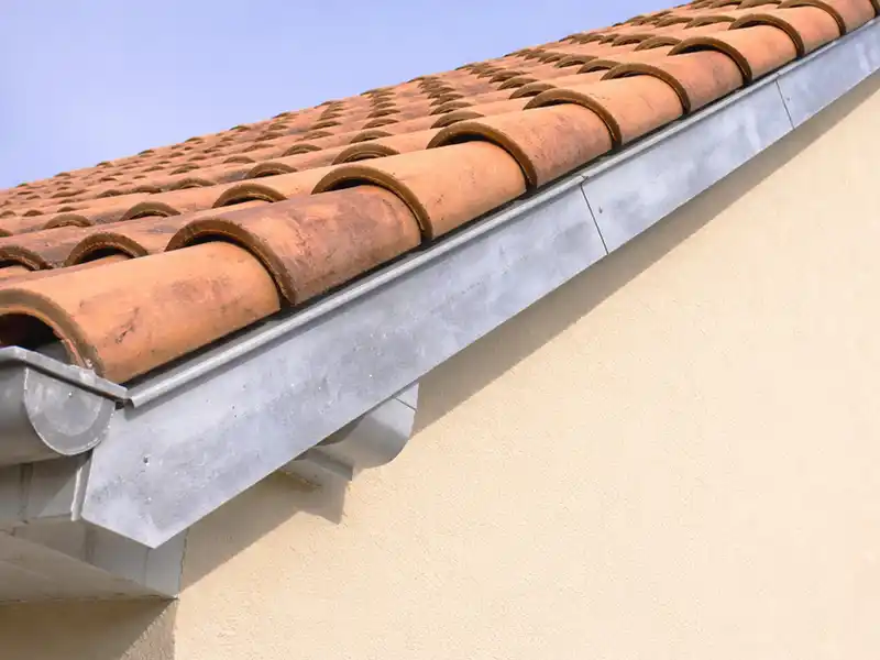 Bande de rive - Zinguerie - Couverture 34 Gros plan sur le toit d'une maison avec des tuiles courbes en terre cuite et une gouttière en métal le long du bord. Le mur est de couleur claire et le ciel est clair et bleu en arrière-plan.