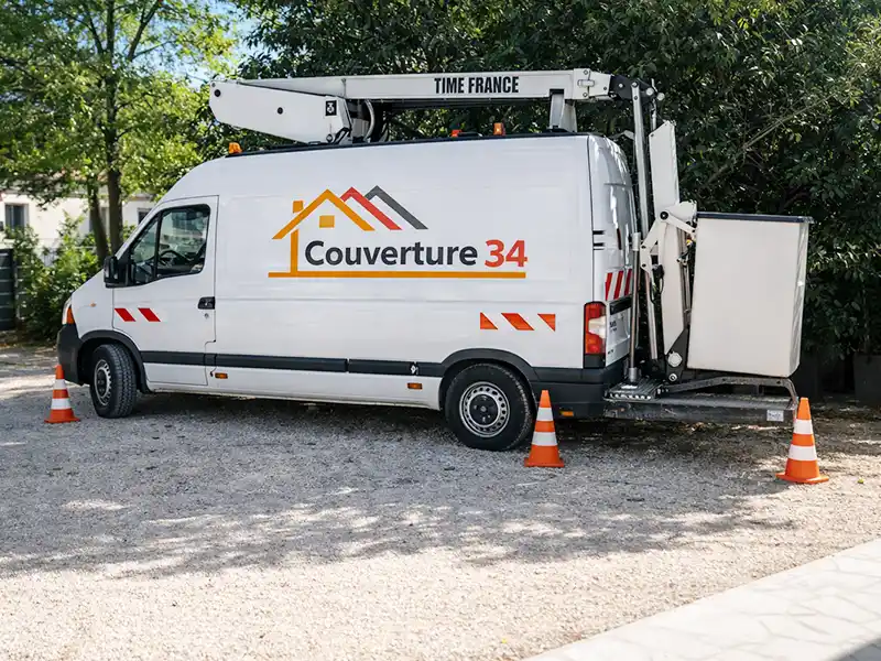 Camion nacelle Couverture 34 - Hérault Une Camion nacelle Couverture 34 blanche avec un logo sur le toit est garée sur du gravier. La camionnette dispose d'une plate-forme de travail surélevée et est entourée de cônes de sécurité orange, avec des arbres en arrière-plan.