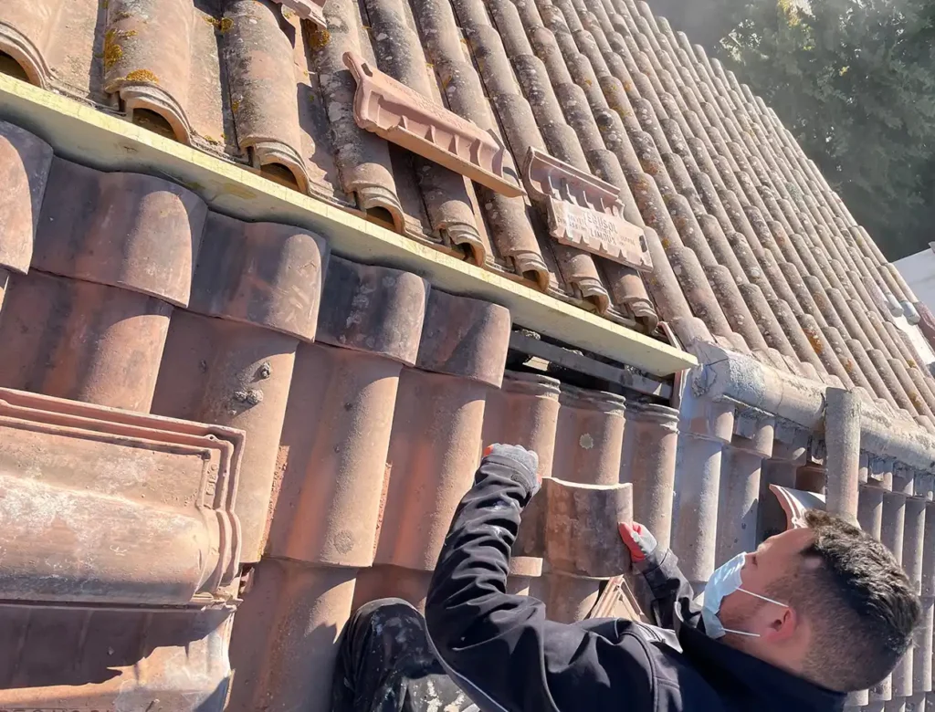 Une personne portant un masque facial répare un toit de tuiles, agenouillée parmi plusieurs tuiles enlevées par une journée ensoleillée, tout en tenant une tuile, en se concentrant sur le changement de faîtage.