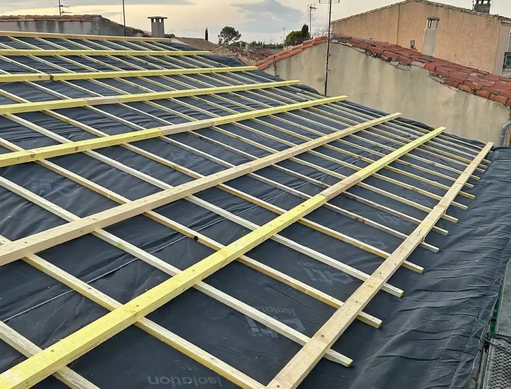 Création de toiture neuve dans le 34 Toit en construction illustrant la création de toiture neuve, avec des lattes de bois disposées en grille sur un matériau d'étanchéité noir, prêtes à recevoir les tuiles, et les toits voisins visibles à l'arrière-plan.