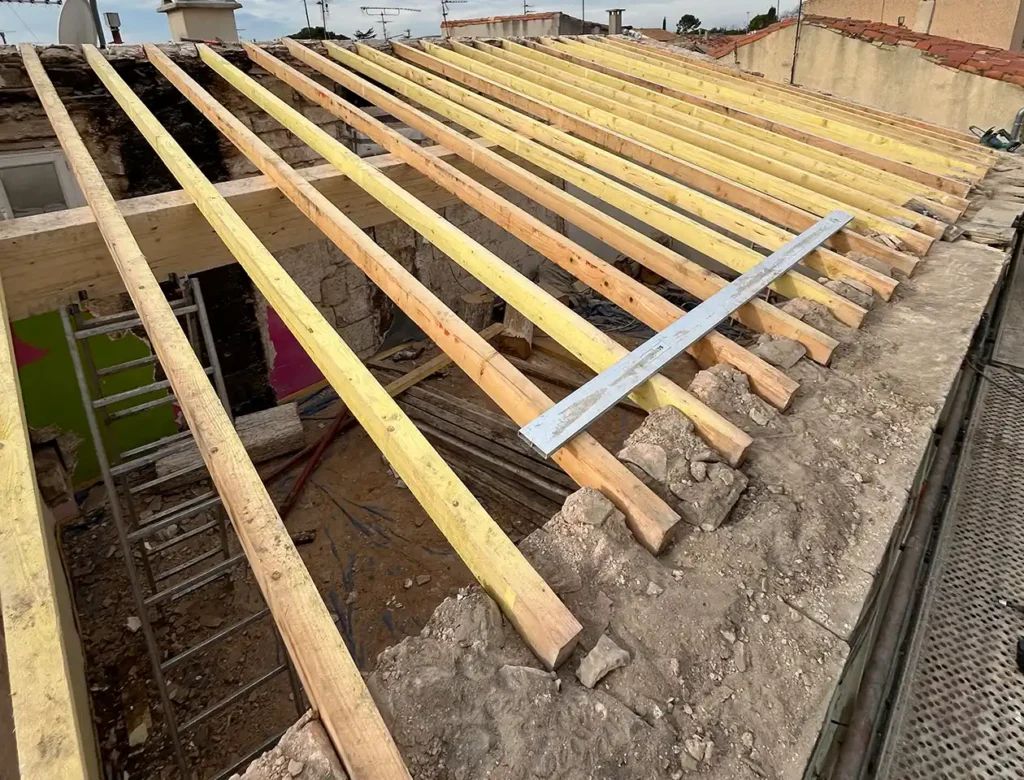 Des poutres en bois sont installées pour former la charpente d'un toit en pente sur un bâtiment en cours de création d'une nouvelle toiture. Des matériaux de construction et des outils sont visibles sous les poutres.