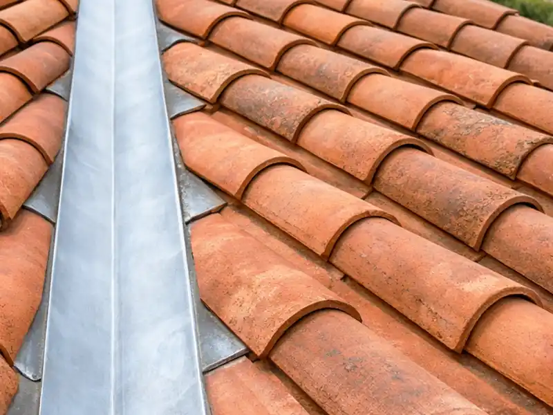 Evacuation des eaux pluviales zinguerie - Couverture 34 Vue rapprochée d'une toiture en tuiles de terre cuite avec une noue métallique pour l'évacuation des eaux pluviales, passant entre deux sections inclinées et mettant en valeur le motif et la texture des tuiles d'argile courbes.