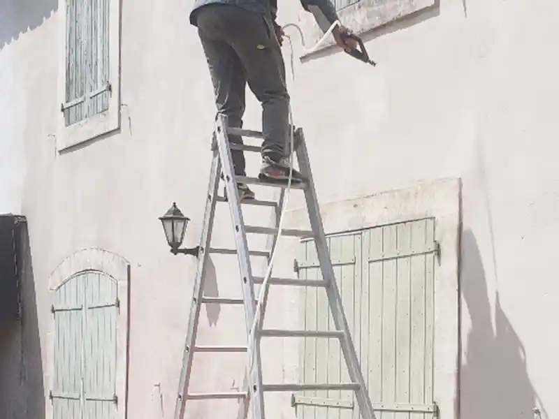 Nettoyage de façade - Couverture 34 Une personne se tient en haut d'une grande échelle, effectuant un nettoyage de façade tout en peignant ou en réparant une fenêtre sur le mur extérieur d'un bâtiment de couleur claire avec des fenêtres à volets et une lanterne murale.