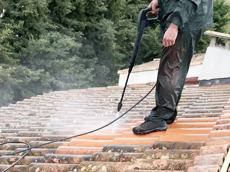 Nettoyage de toiture - Couverture 34 Une personne portant des vêtements imperméables utilise un nettoyeur à pression pour le nettoyage de toiture, enlevant la mousse et la saleté des tuiles d'un toit en pente, avec des arbres visibles à l'arrière-plan.