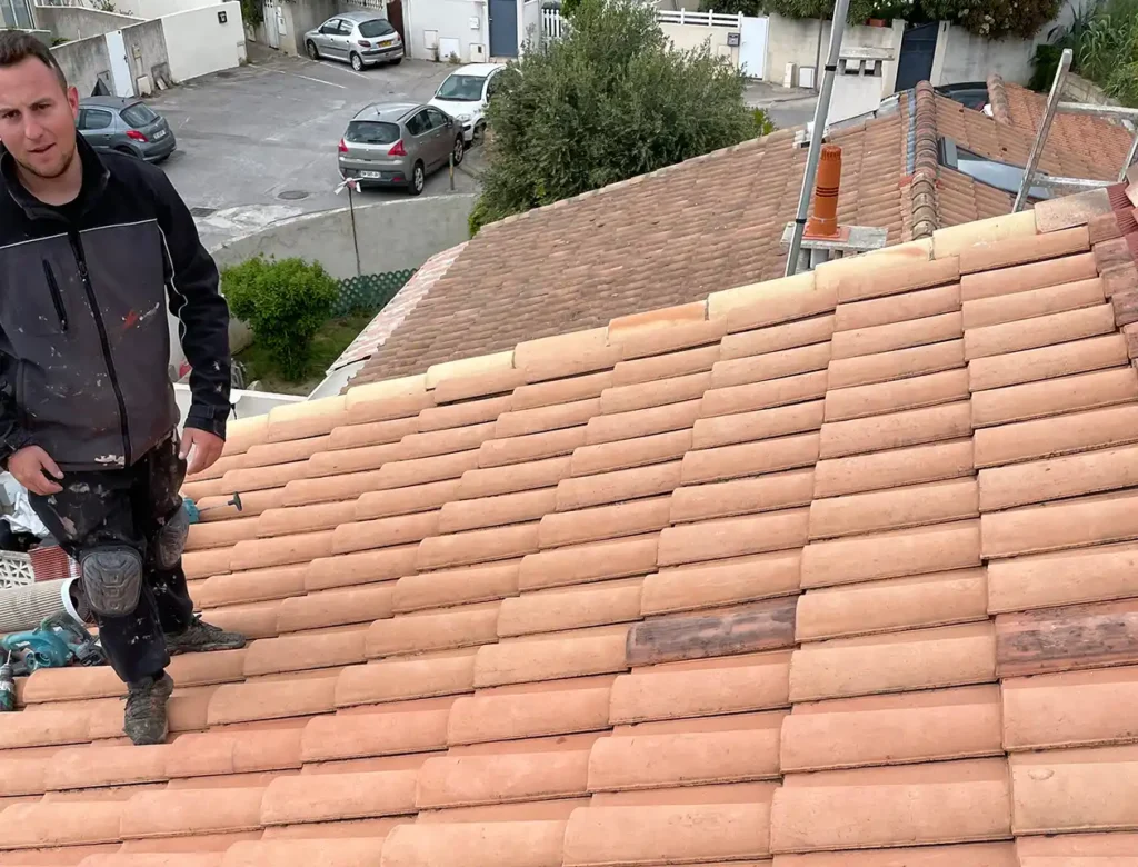 Nettoyage de toiture complet (1) Un homme en vêtements de travail se tient sur un toit incliné en tuiles de terre cuite, effectuant un nettoyage de toit, avec un échafaudage et un quartier résidentiel avec des voitures garées visibles à l'arrière-plan.