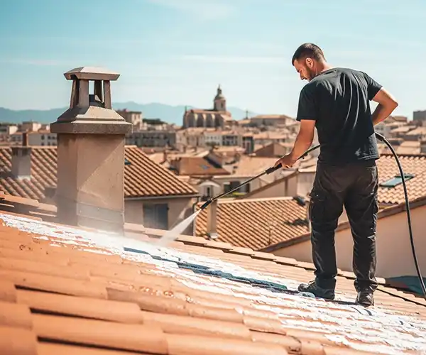 Nettoyage et démoussage de toiture à Lunel - Couverture 34 Un homme se tient sur un toit en tuiles, effectuant un nettoyage et démoussage de toiture avec un nettoyeur à pression pour nettoyer les graffitis. Des bâtiments et une église apparaissent à l'arrière-plan sous un ciel clair.