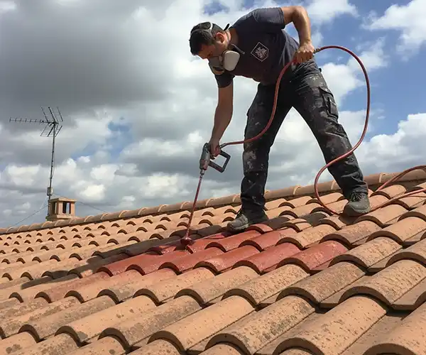 Peinture de toit à Lunel - Couverture 34 Une personne se tient sur un toit en tuiles, portant un masque et appliquant de la peinture de toit à l'aide d'un pistolet, ce qui rend les tuiles rouges. Le ciel est partiellement nuageux et une antenne est visible à l'arrière-plan.
