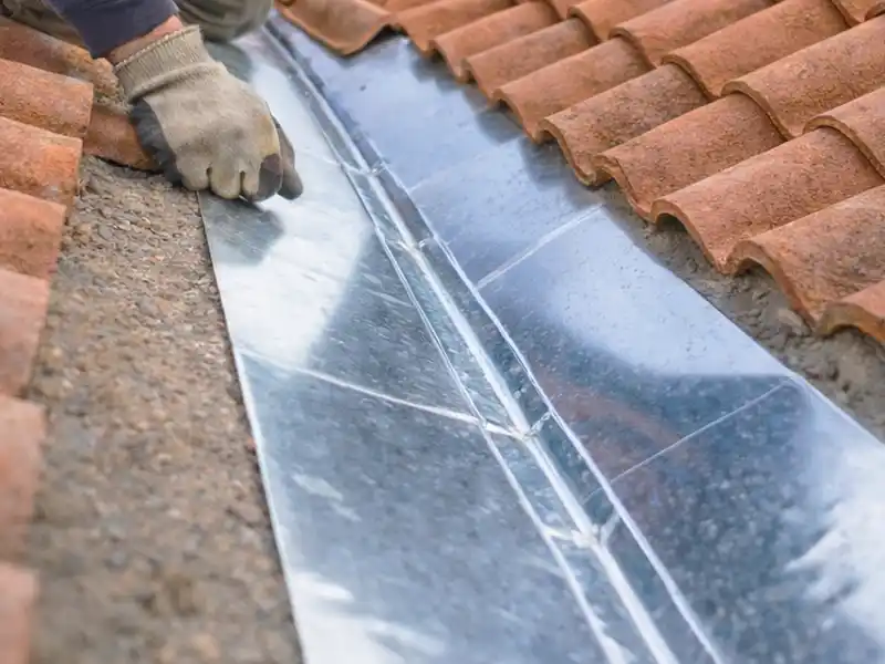 Une personne portant un gant installe un solin de toit en m&eacute;tal pendant la pose d'une noue, le pla&ccedil;ant entre les tuiles en argile rouge et la surface rugueuse des bardeaux sur un toit en pente.