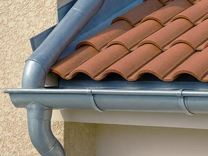 Pose et nettoyage de gouttière dans le Gard - Rénovation Sud Vue rapprochée d'un toit de maison avec des tuiles en terre cuite rouge et un système de gouttière en métal argenté fixé au bord du toit et à la descente d'eau, sur un mur extérieur beige.