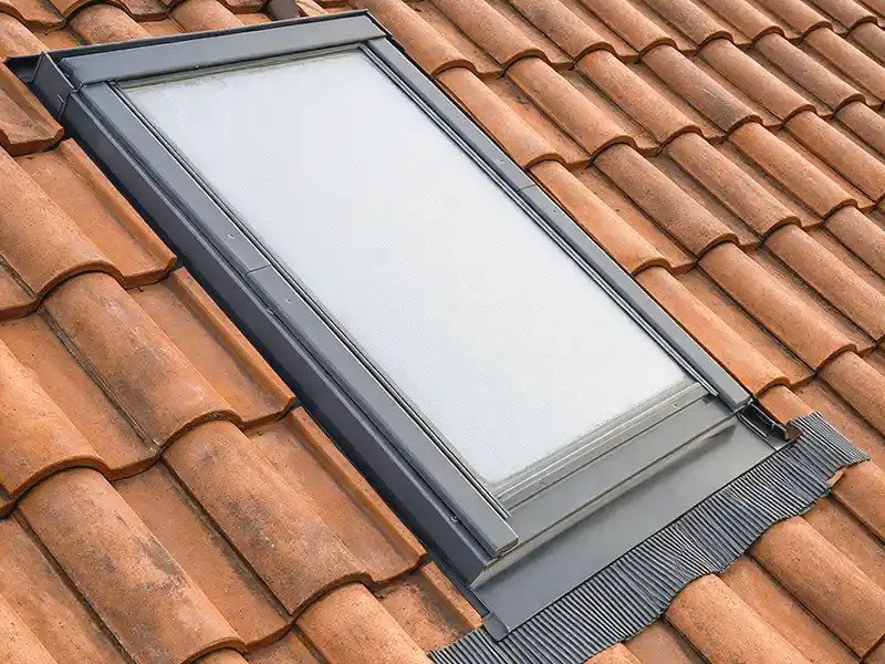 Pose fenêtre de toit à Nimes et dans le Gard - Rénovation Sud Une fenêtre de toit rectangulaire avec un cadre noir est installée sur un toit avec des tuiles en terre cuite brun-rouge. Cette fenêtre de toit est dotée d'un verre légèrement dépoli et d'un solin d'étanchéité à la base.