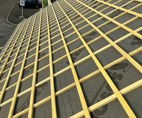 Une toiture en cours de construction lors d'une r&eacute;novation de toiture, avec une grille de lattes en bois r&eacute;guli&egrave;rement espac&eacute;es sur une sous-couche noire, vue d'en haut et de biais. Une rue avec des voitures gar&eacute;es et un panneau de signalisation est visible &agrave; l'arri&egrave;re-plan.