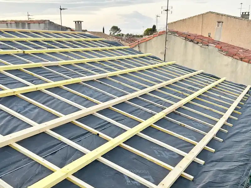 Rénovation du toit - Couverture 34 Toit en pente en cours de construction lors d'une rénovation du toit, recouvert d'une membrane d'étanchéité noire et de lattes de bois parallèles disposées en grille, avec les toits voisins et les antennes visibles à l'arrière-plan.