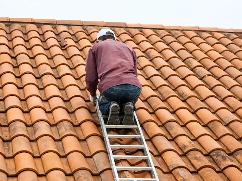 Une personne portant une casquette blanche, une chemise marron et un jean s'agenouille sur un toit en tuiles lors d'un diagnostic de toiture, en regardant vers le haut et en s'appuyant sur une &eacute;chelle en aluminium.