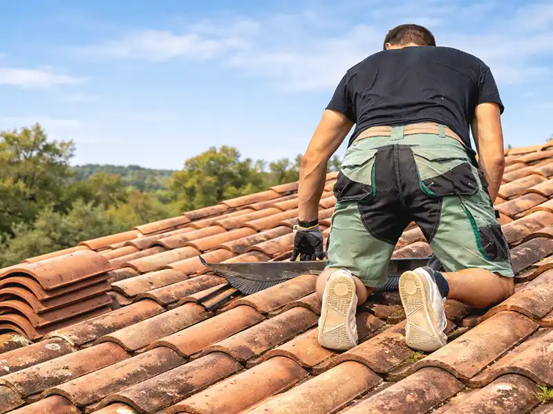 Réparation de tuiles dans le déparement Hérault - Couverture 34 Une personne portant des gants de travail et un short est agenouillée sur un toit de tuiles rouges, réparant ou inspectant les tuiles sous un ciel bleu ensoleillé - un exemple de Réparation de tuiles dans le département de l'Hérault avec des arbres en arrière-plan.