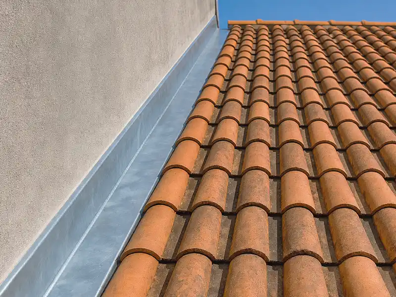 Travaux de zinguerie et étancheité dans le Gard - Rénovation Sud Vue rapprochée d'un toit en pente avec des tuiles en terre cuite orange, bordé d'un solin métallique gris à la jonction du toit et d'un mur en stuc, sous un ciel bleu clair.